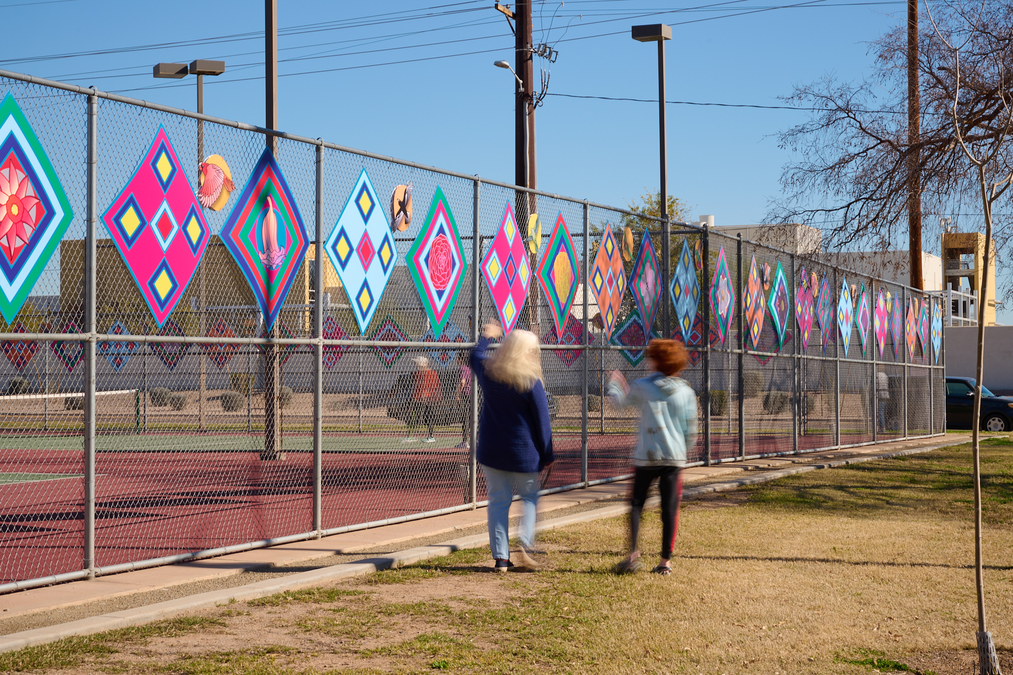 Image of a fence with bright, multicolored diamond-shaped panels hanging in a row across the top. People walking in front and looking up at the art.