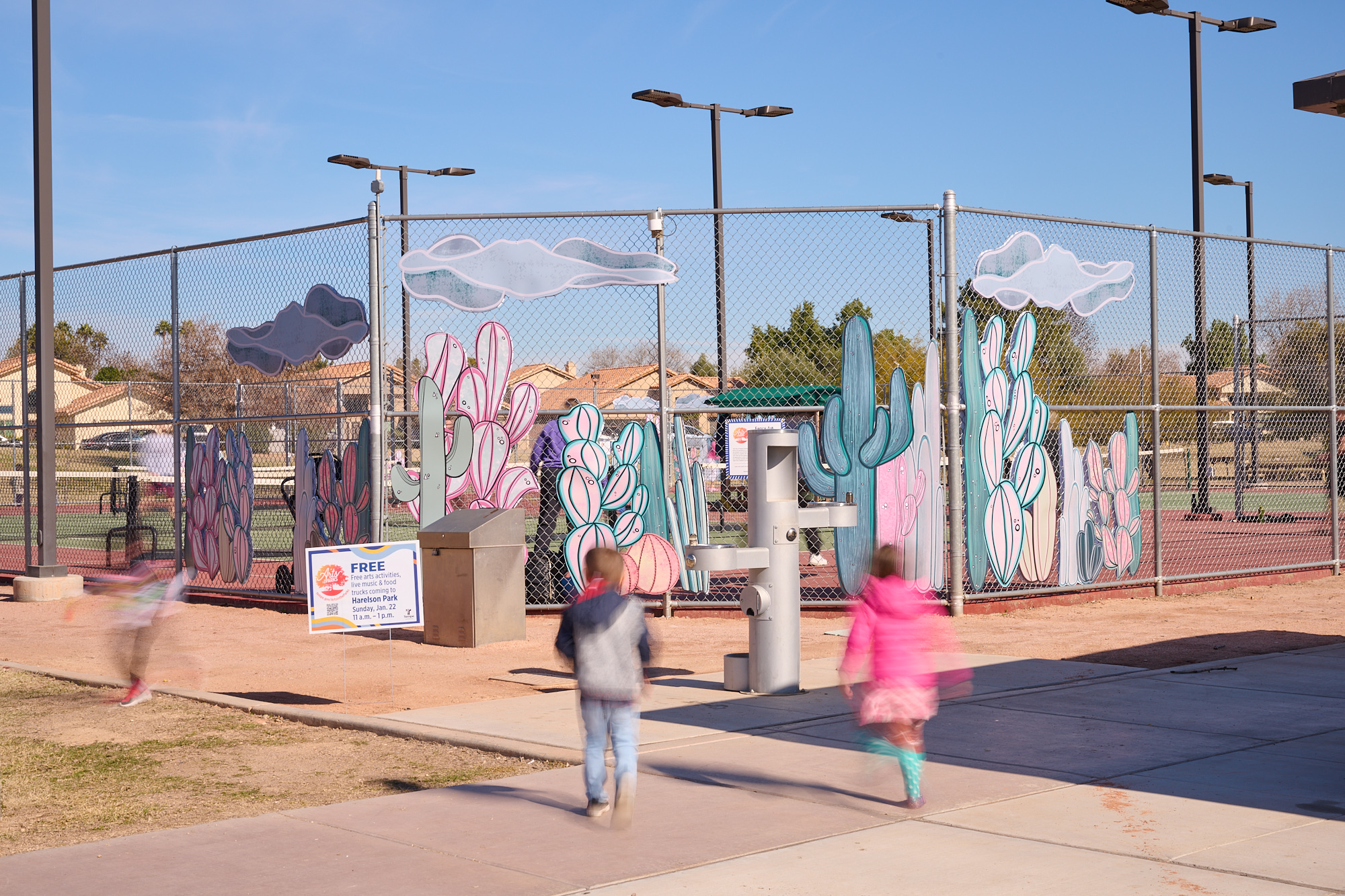 Image of a fence with two children in front. The fence has art of pastel-colored cacti and clouds attached.