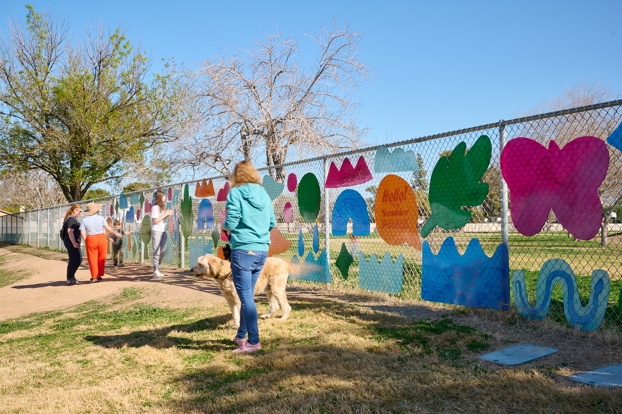 Image of a fence with people walking in front. Along the fence are bright-colored shapes of different things including a pink butterfly and a green leaf.