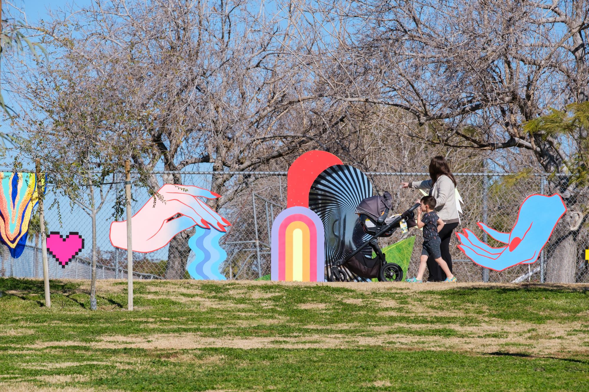 Photo of a fence with bright graphic art installed. Woman with a baby stroller walking passed on the street. The fence has a reflective rounded mirror and a large pink hand with watery drips flowing down.