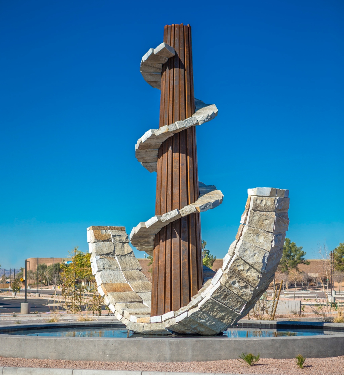 In the middle of the image is a thick, brown, metal pole which extends high into the air. Curved beneath the pole is a wide stone curve which reaches halfway up the pole. Running along the whole metal pole is a stone spiral which juts out like a walkway.