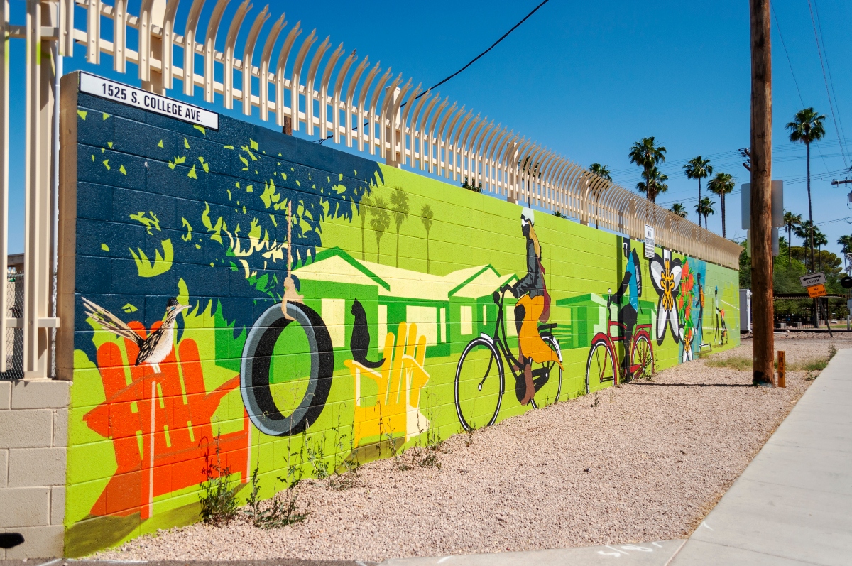 This picture shows a wall that is painted with a bright, colorful mural.bThe mural shows a neighborhood scene with houses in the background. In the foreground are people riding bikes, a chair with a cat, and a tire swing on a tree.