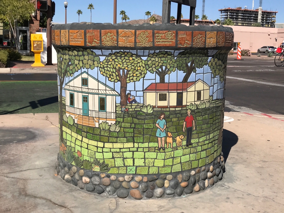 A cylindrical standpipe is in the foreground. The standpipe is covered in a colorful mosaic made out of tiles and rocks. The mosaic depicts two houses in a green field with trees in the background. In the foreground of the mosaic are a white man and woman walking a dog.