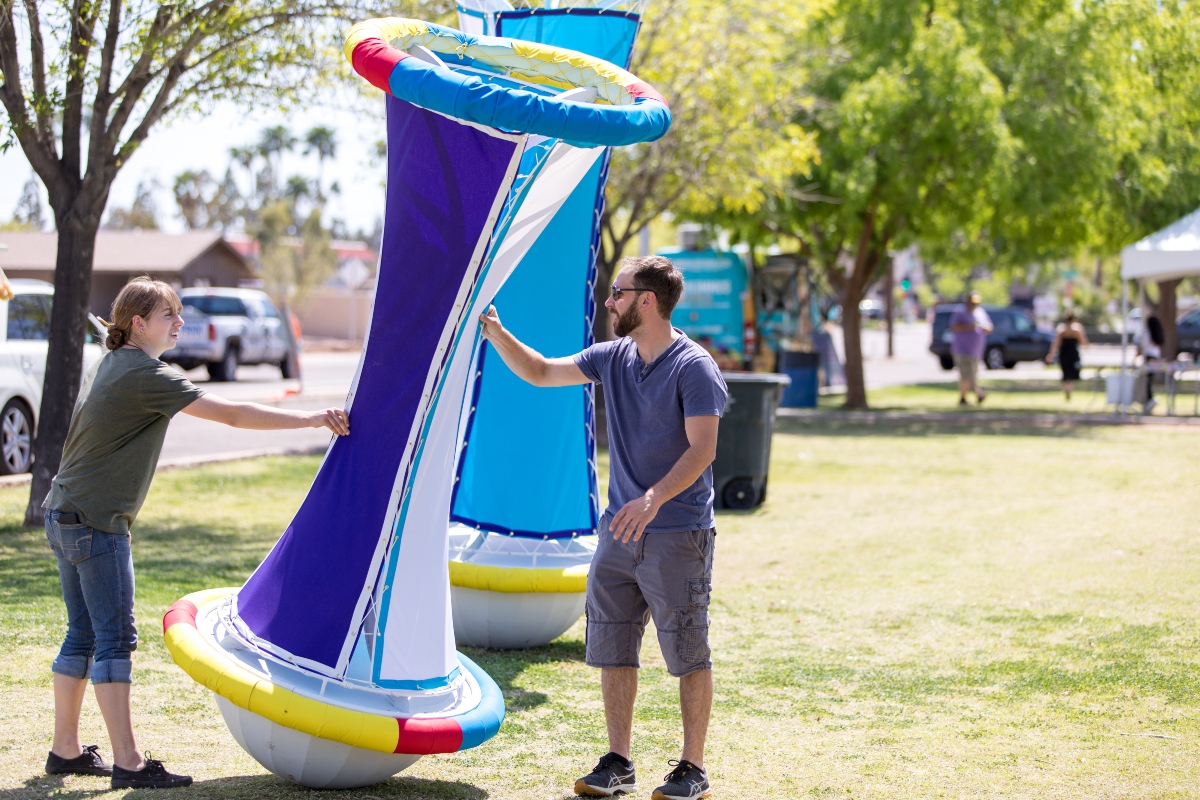 Two people stand in a park on a sunny day in while touching a tall sculpture. The sculpture is blue and white. It has a rounded top and bottom with a tube-like middle section. It stands upright.