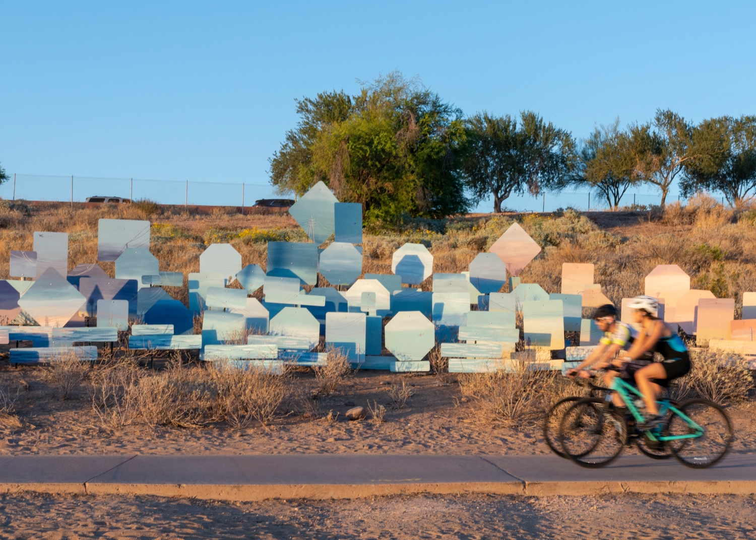 A dry field with a path in front. Two bikers in the far right frame are blurry as they ride. In the field are over 20 kinds of traffic signs. The signs are painted in varying shades of blues, whites, and peach. They look like the sky during the day, sunrise, and sunset.