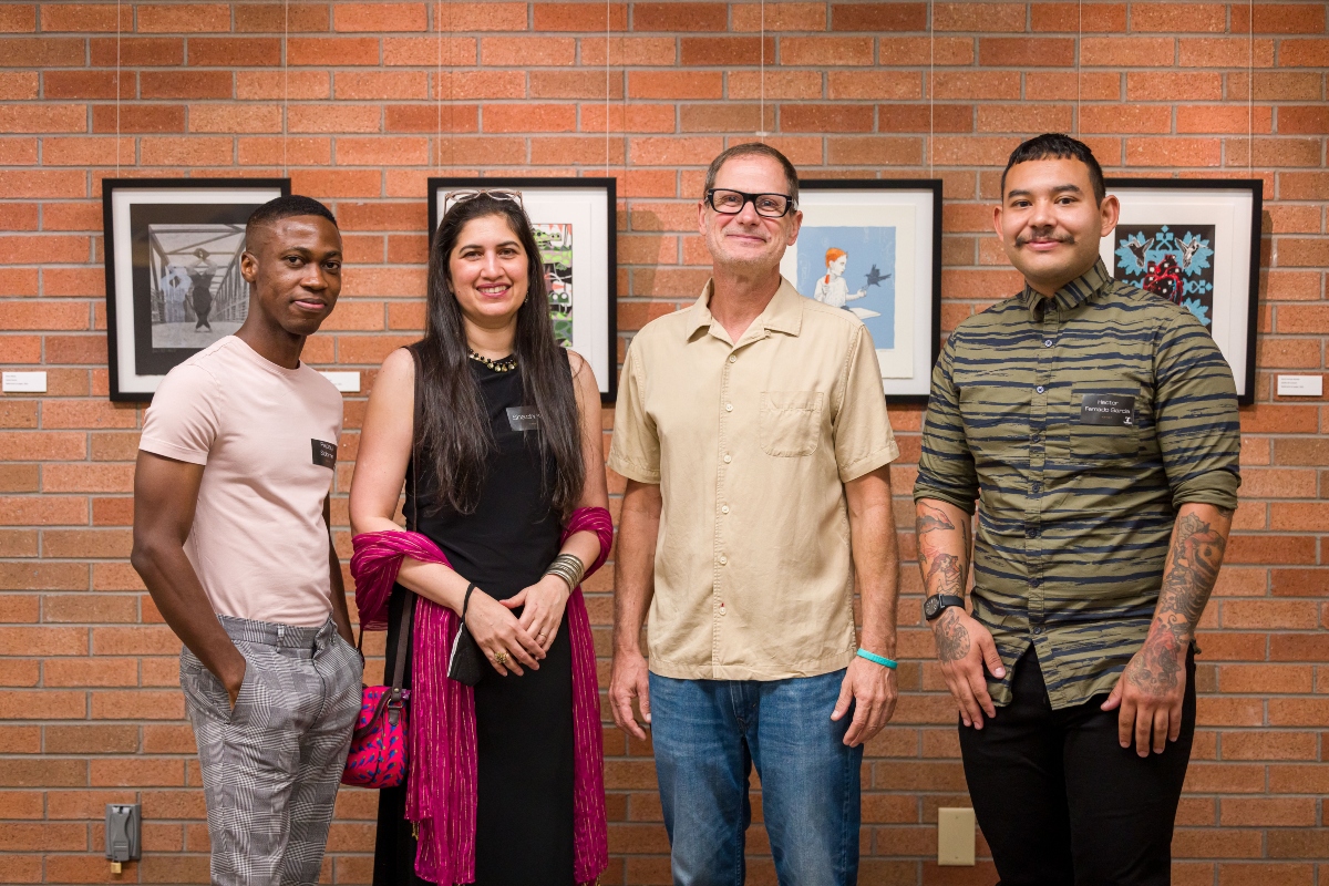 A black man, Indian woman, white man, and Hispanic man stand in front of a brick wall. On the wall are hanging four art prints.