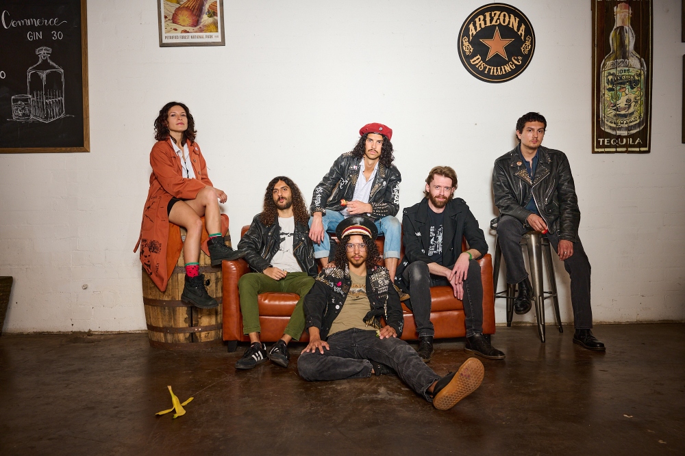 One woman and 5 men stand and sit around an orange couch.