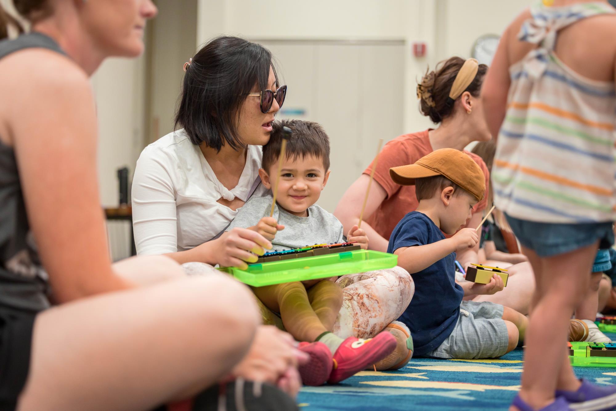 A woman holds a young boy on her lap as they play a xylophone 