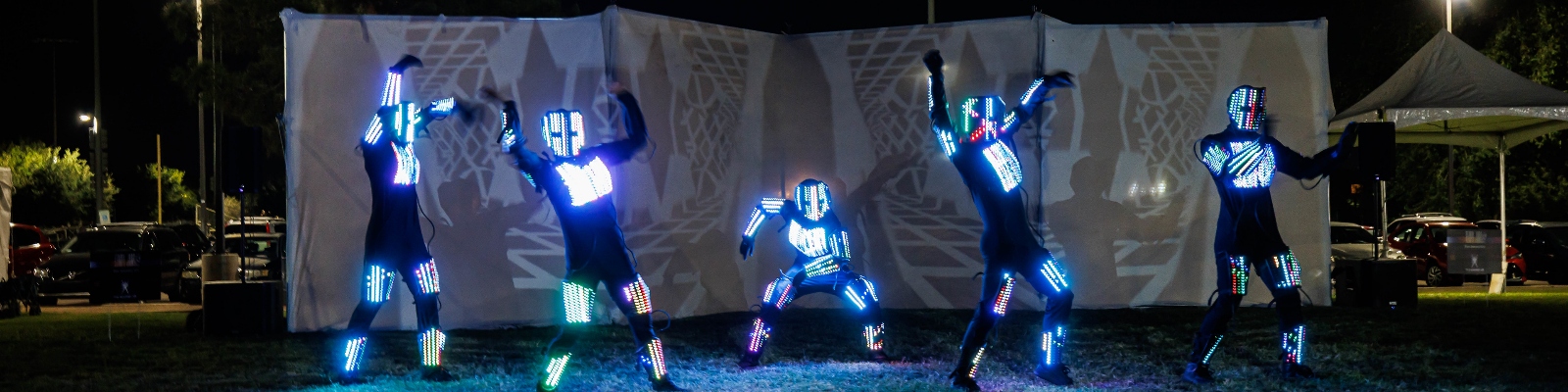 The Sacred Gs perform in LED light suits in front of a geometric projection.