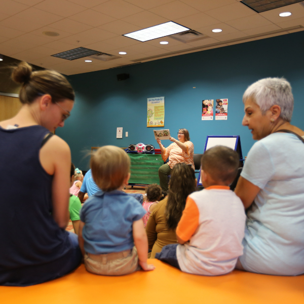 Two little children sit next to their caregivers while listening to someone read a story at the library.